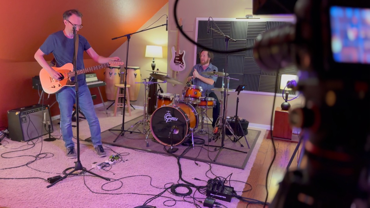 Live session: guitarist and drummer performing, Blackmagic camera and monitor visible in foreground
