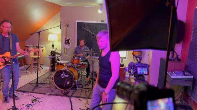 Travis operating camera during live session: guitarist, drummer, softbox, Blackmagic monitor